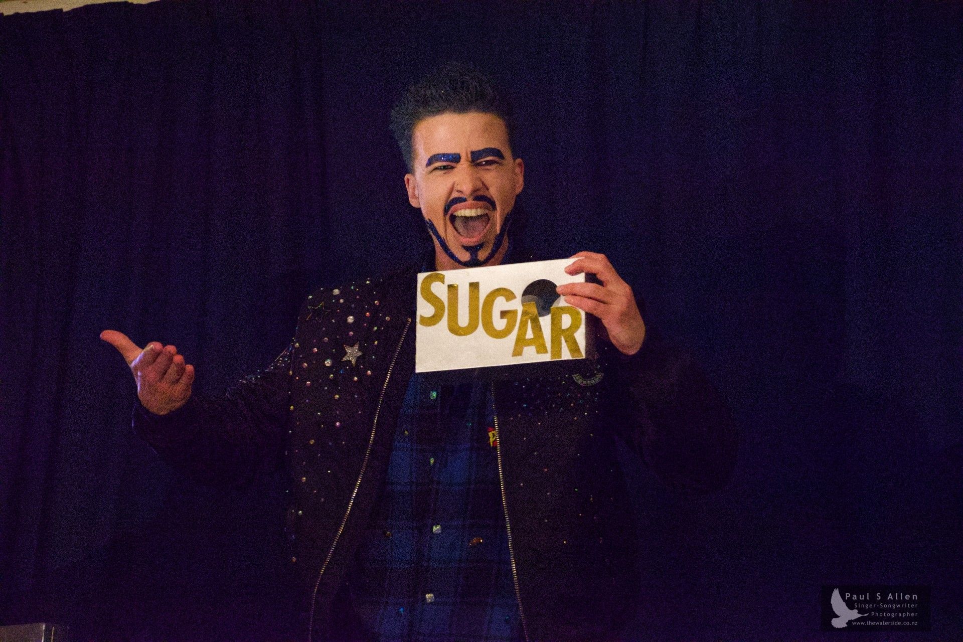 Hayes pictured in drag with thick brows and a beard. He’s wearing a dark jacket covered in stars, and holds a card that says ‘SUGAR’ in large gold letters.