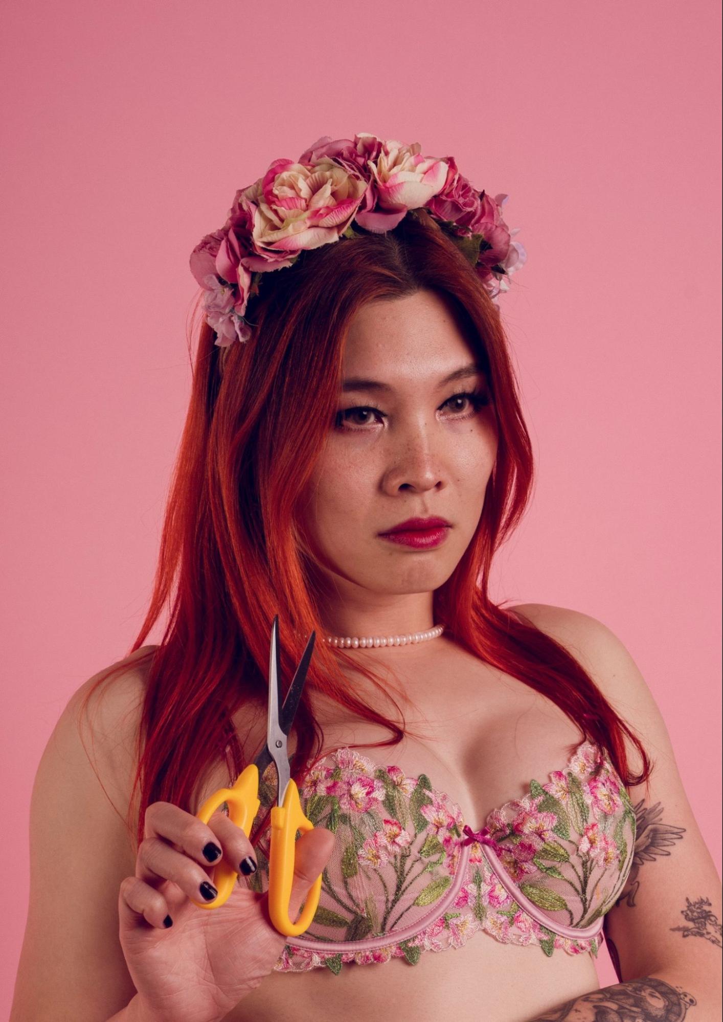 Lily Catastrophe sits in an embroidered bra, wearing a floral headband, holding a pair of open scissors. She has bright red long hair, red lips, and is wearing a pearl necklace.